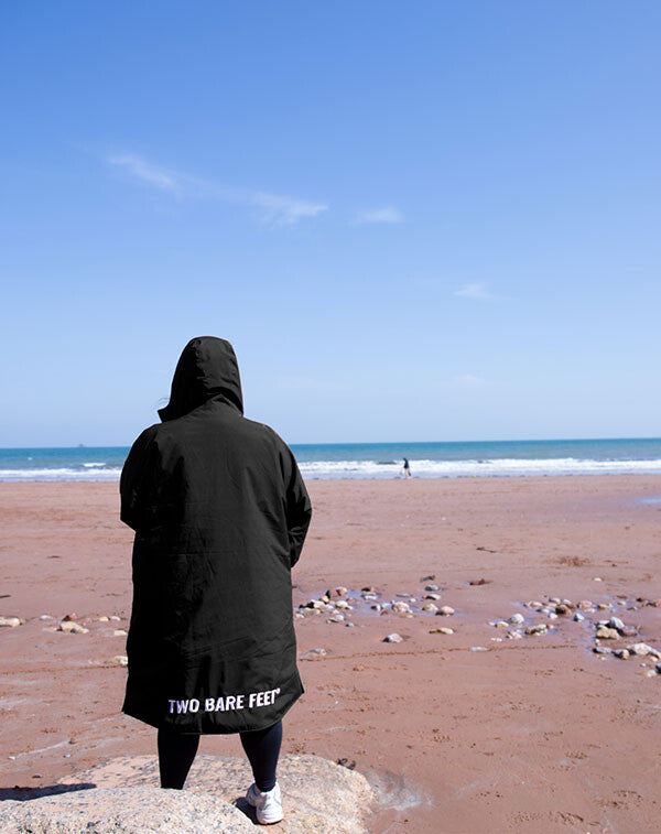 Person wearing a black hooded changing robe with 'Two Bare Feet' branding on a beach.