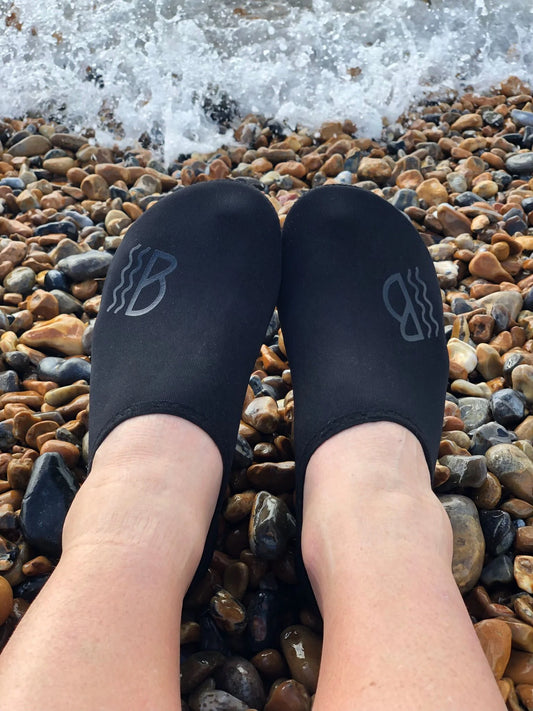 Black neoprene water shoes on feet standing on pebbles with ocean waves in the background
