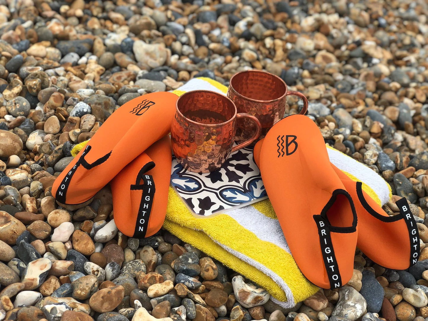 Orange neoprene water shoes with 'Brighton' branding on a pebbly surface with two copper mugs.